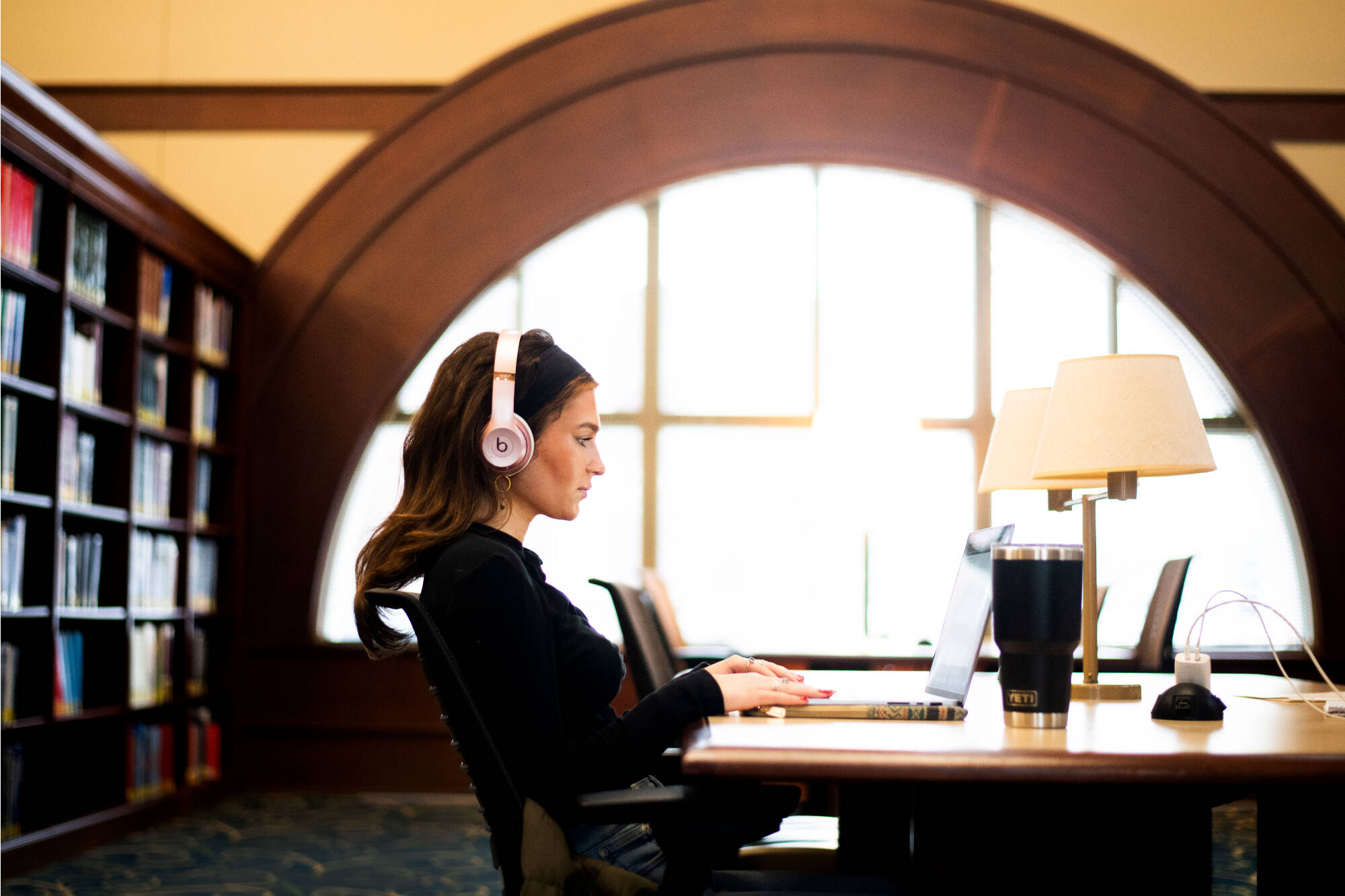 student sitting with headphones on at a library table in front of archway windows